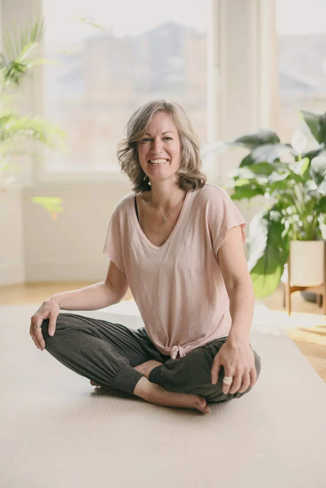 Nicki sitting cross-legged on a mat in a bright studio with plants, smiling