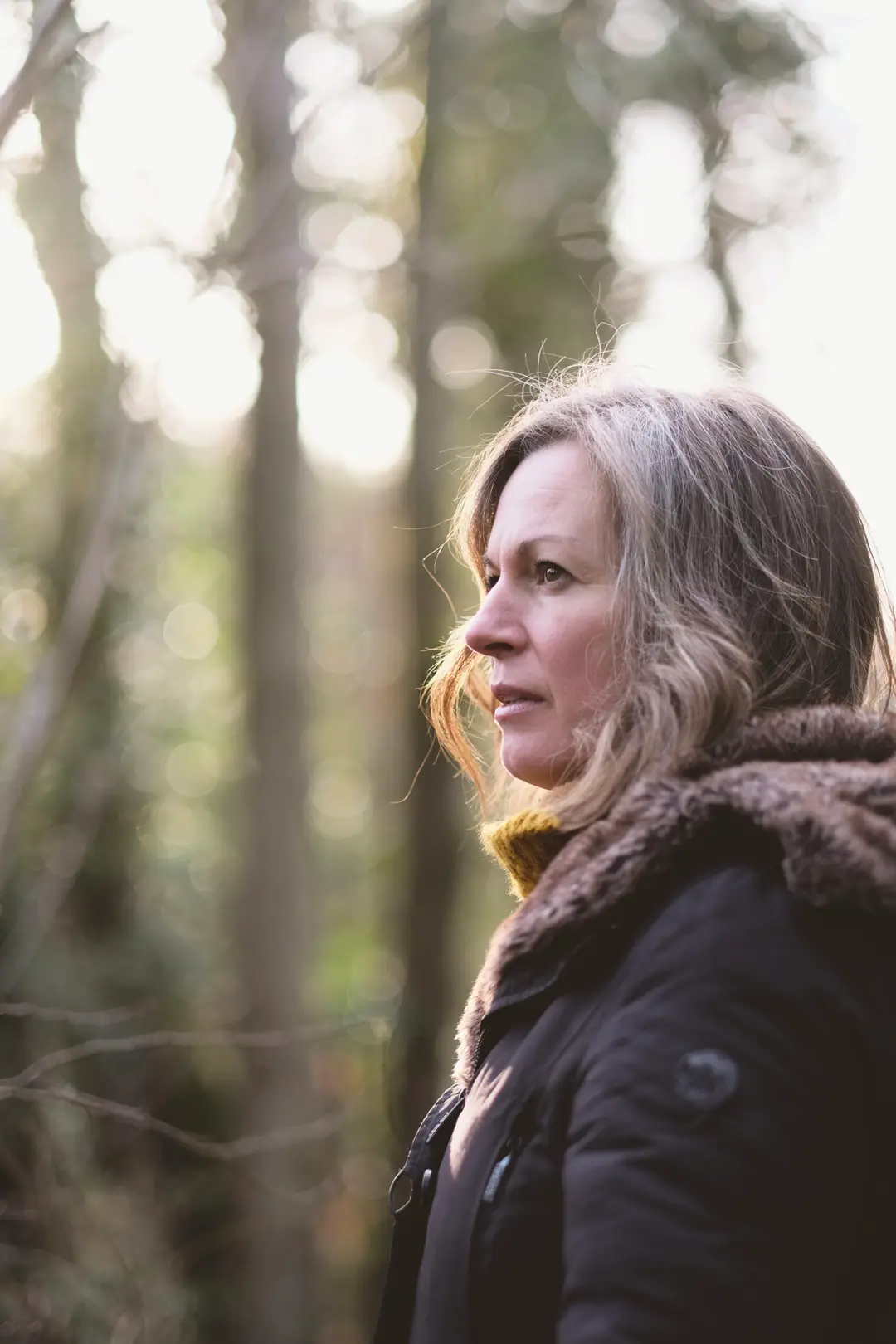 Nicki in a winter coat among tall trees, looking into the distance